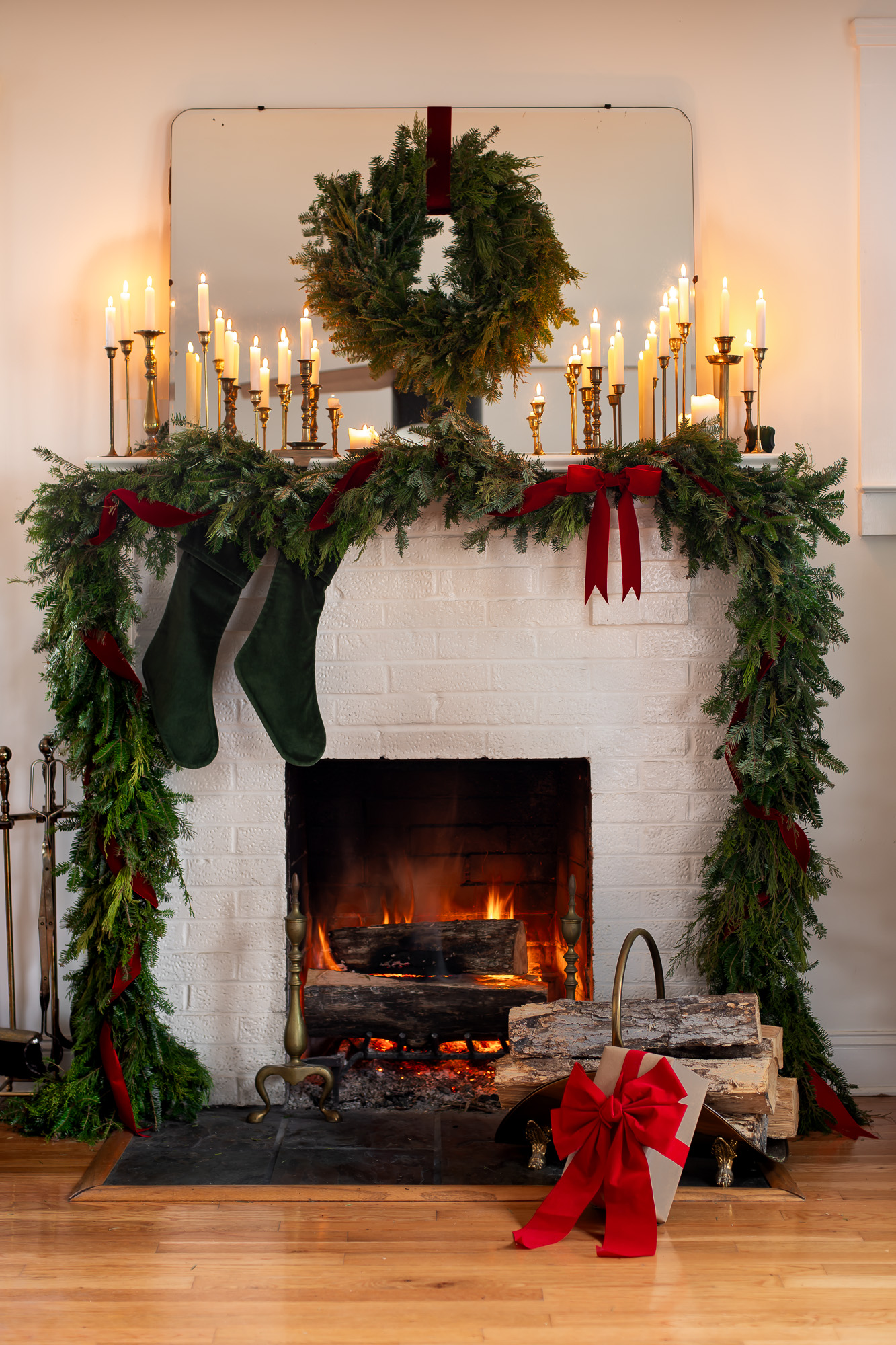 Old World Christmas Mantle with evergreen garland trimmed with red ribbon, green velvet stockings hanging over a roaring fire and dozens of lit candles in brass candlestick holders with a fresh evergreen wreath hanging by a red velvet ribbon.