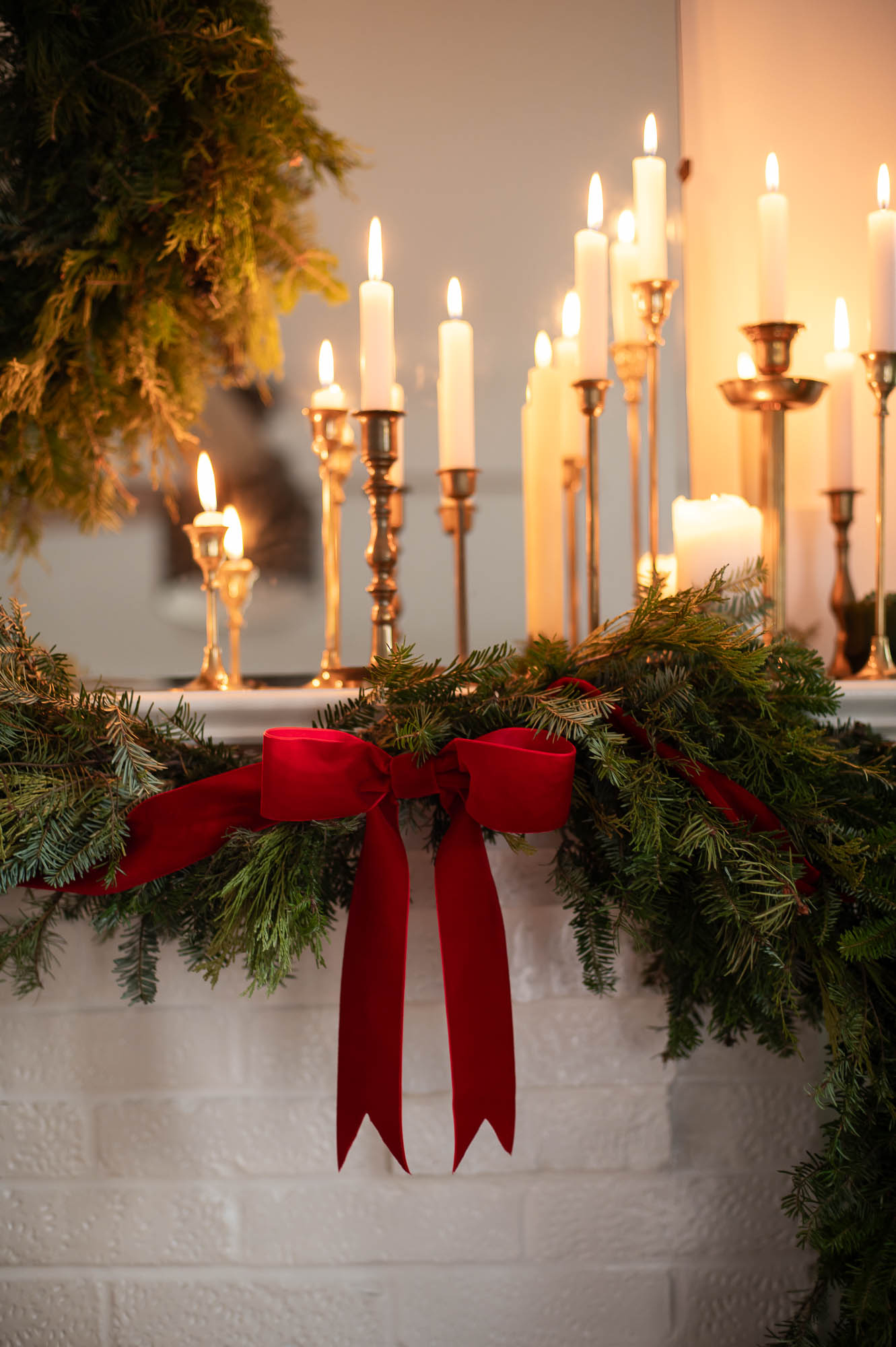a red velvet ribbon is woven through a fresh evergreen garland and tied in a bow. Candles in brass candlesticks are lit on the mantle above.