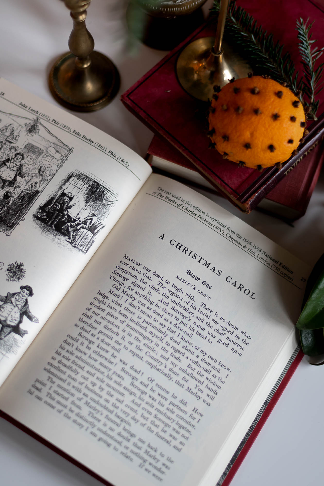 an illustrated edition of A Christmas Carol lays open on a table surrounded by vintage book, clove studded oranges and brass candlestick holders.