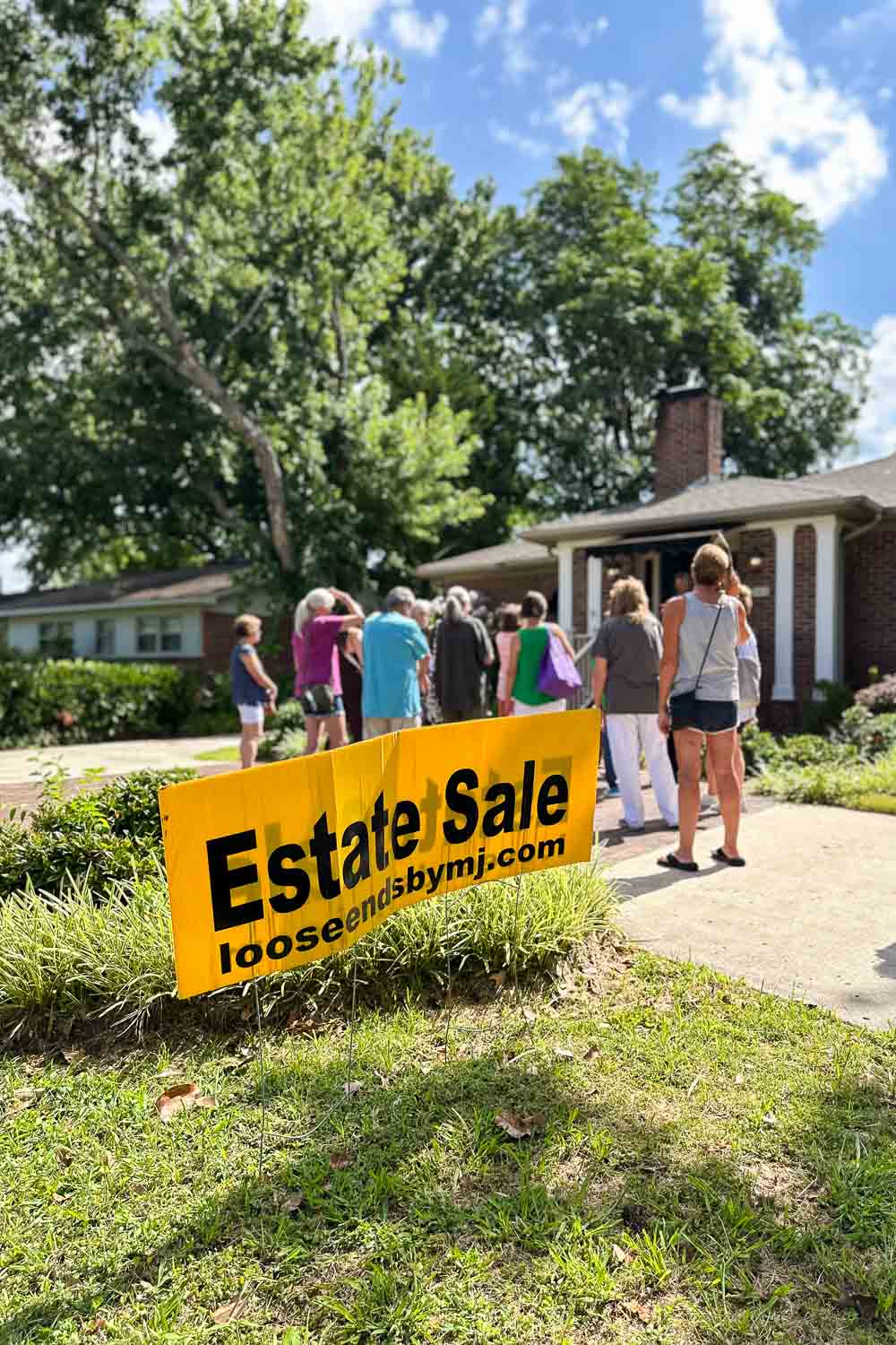 Estate sale signs posted outside suburban house showing directions to weekend sale with a line of people waiting out front