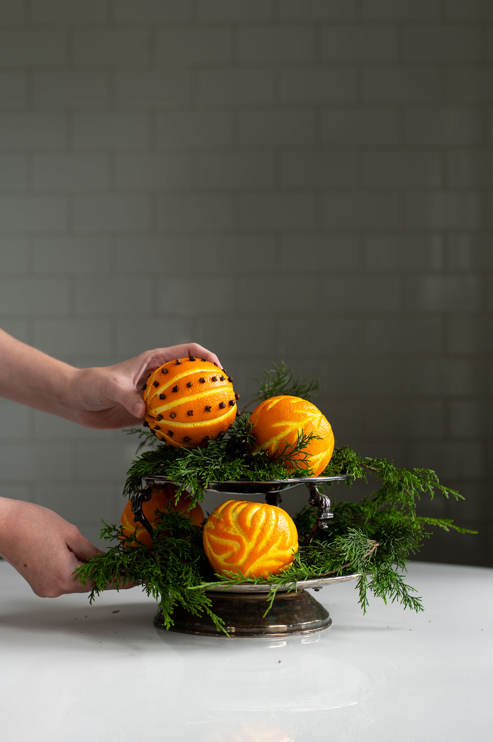 a woman places a orange clove pomander ball onto a silver tray filled with christmas greenery