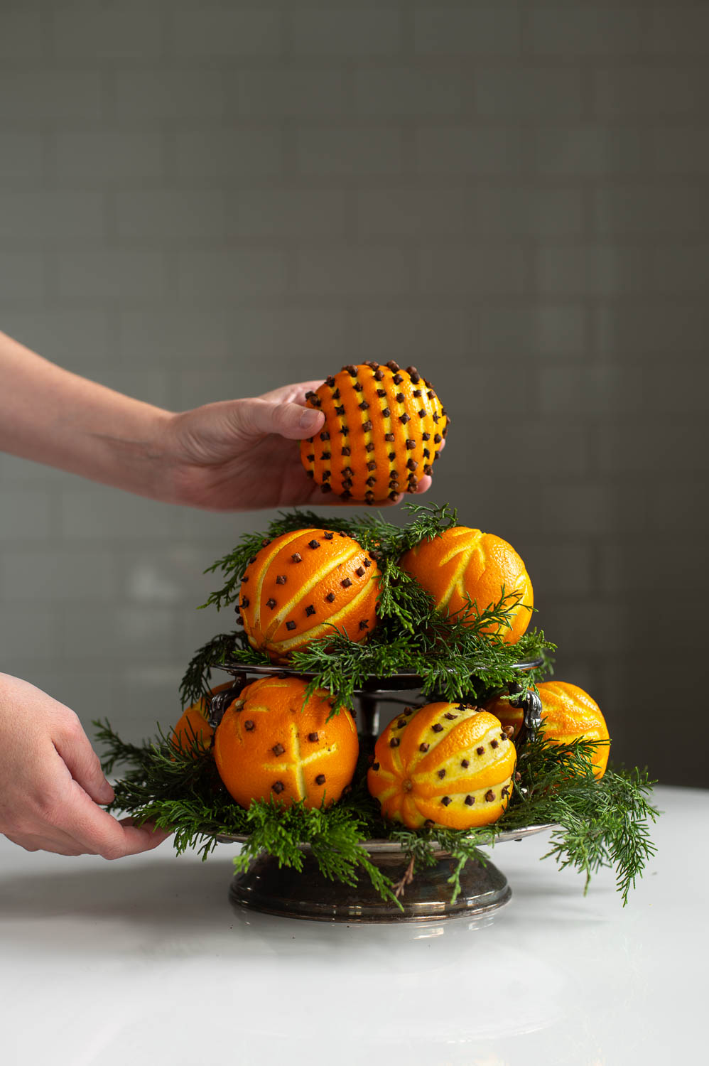 a woman places a orange clove pomander ball onto a silver tray filled with christmas greenery to create a natural christmas decor centerpiece.
