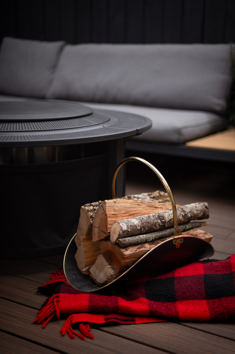 Cozy fire pit party decor with logs stacked in a black and brass metal log holder and plaid blankets – perfect for a fall bonfire night party.
