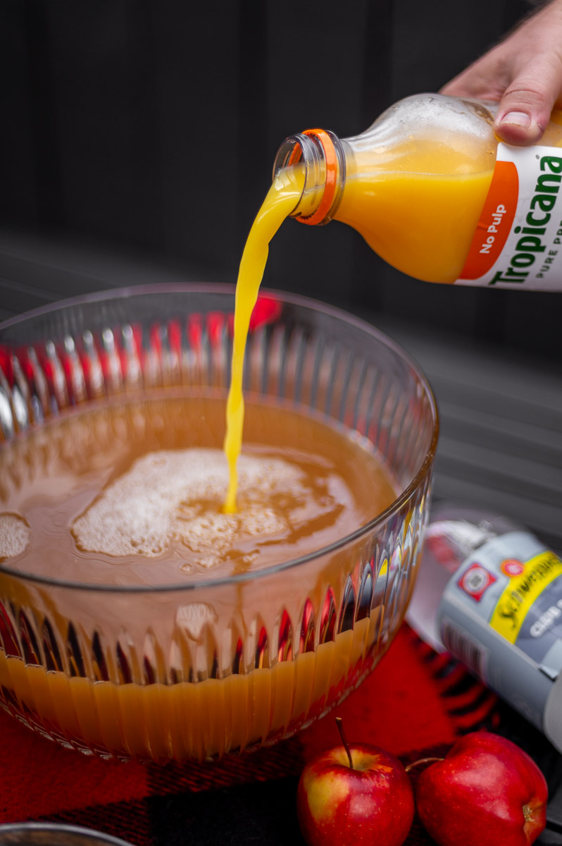 Orange juice being poured into a vintage glass punch bowl to make apple cider punch non alcoholic, ideal for fall punch recipes non alcoholic, harvest punch, and Thanksgiving punch non alcoholic.