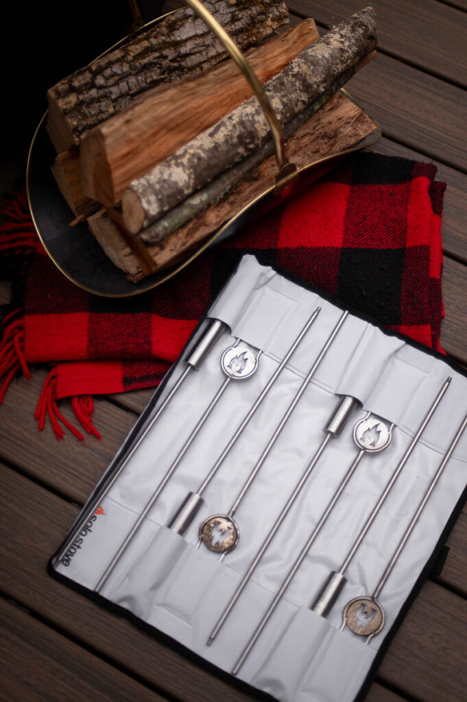 solo stove roasting sticks shown displayed in their storage case opened on wood deck next to a red flannel blanket and rack of firewood.