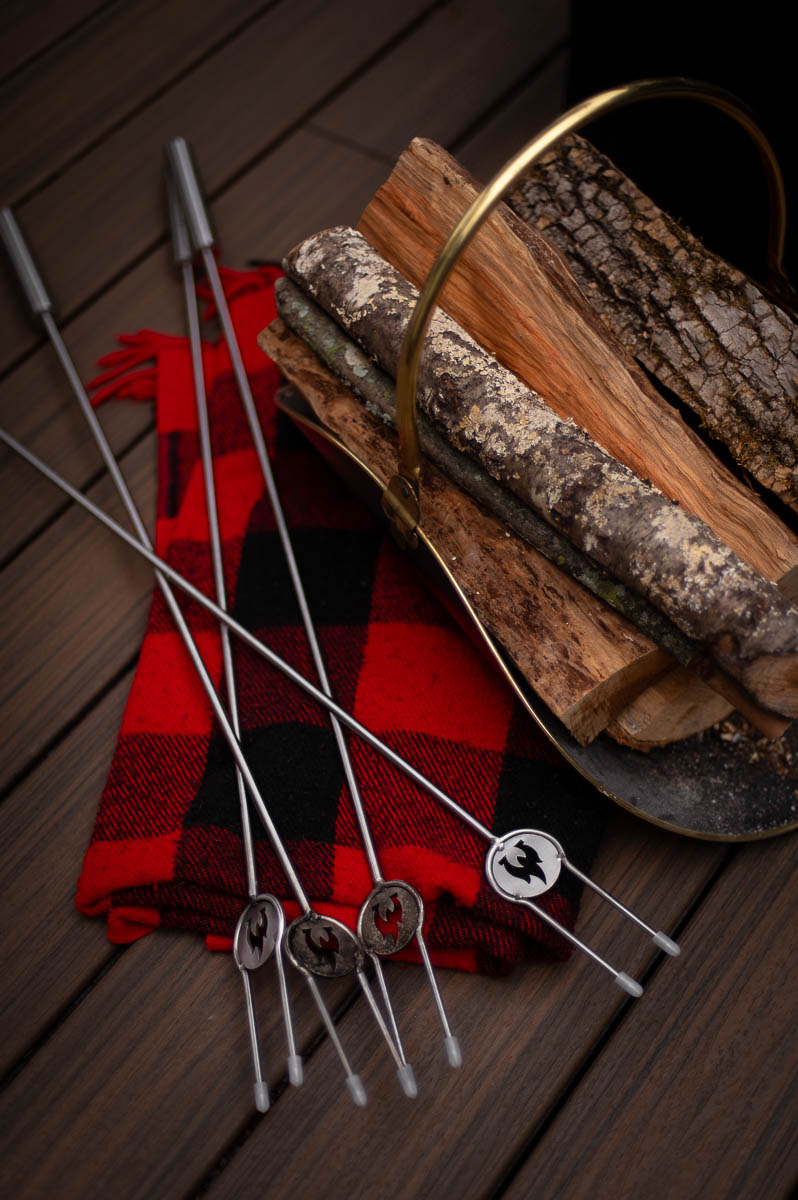 Solo stove fire roasting sticks shown laying on buffalo check blanket on top of a trex composite deck alongside a rack of firewood.