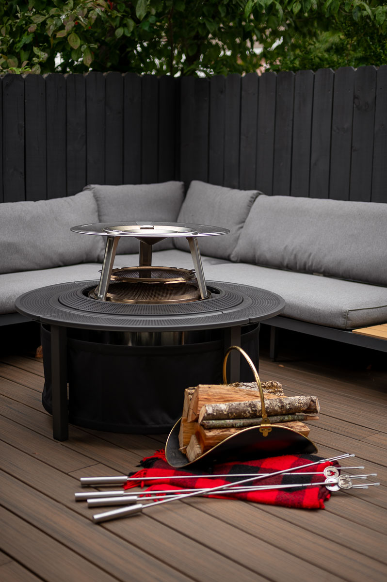 Solo Stove Bonfire set up on a wooden deck with heat deflector, spark shield, fire pit surround, fire roasting sticks next to some cozy seating.