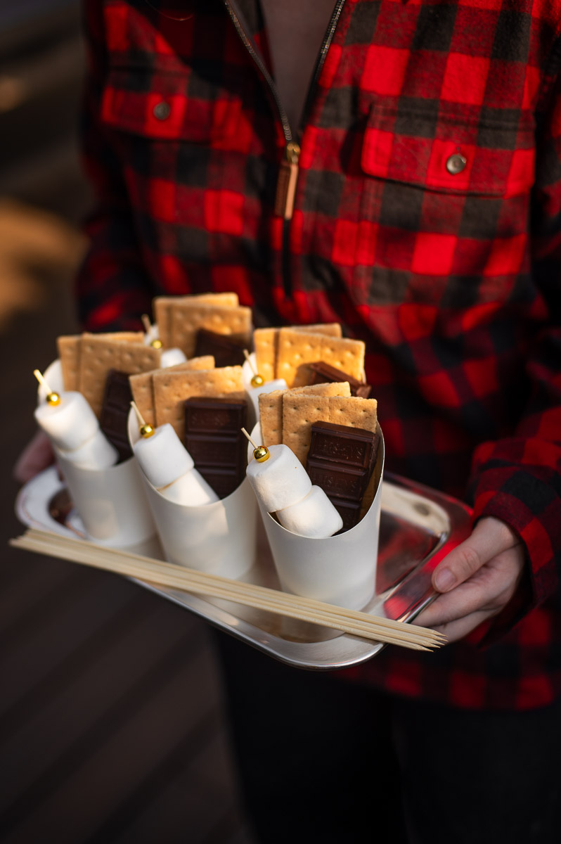 A woman holds a silver tray filled with homemade individual s'mores kits containing marshmallow skewers, chocolate bars and graham crackers at a camping themed party