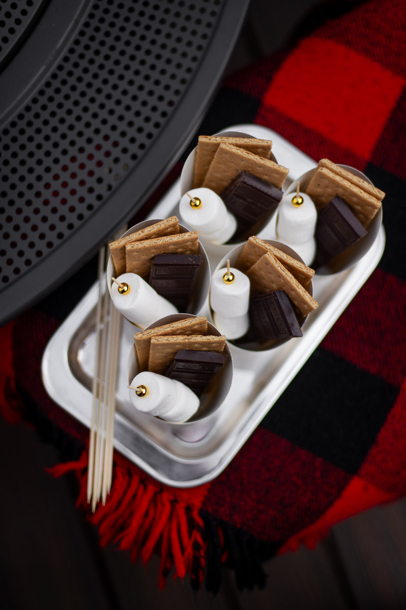 DIY S'mores kits filled with marshmallow skewers, chocolate bars and graham crackers sit on a silver tray on a red and black flannel blanket ready to serve at a bonfire party