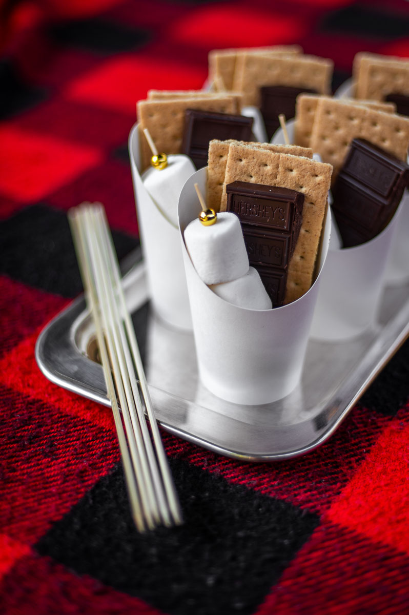 DIY S'mores Cups filled with marshmallow skewers, chocolate bars and graham crackers sit on a silver tray on a red and black flannel blanket ready to serve at a backyard BBQ party