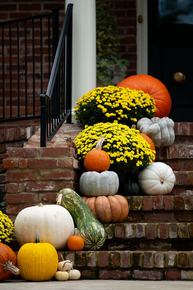 pumpkin decorating on a fall front porch with colorful stacked pumpkins and mums lining a brick staircase.