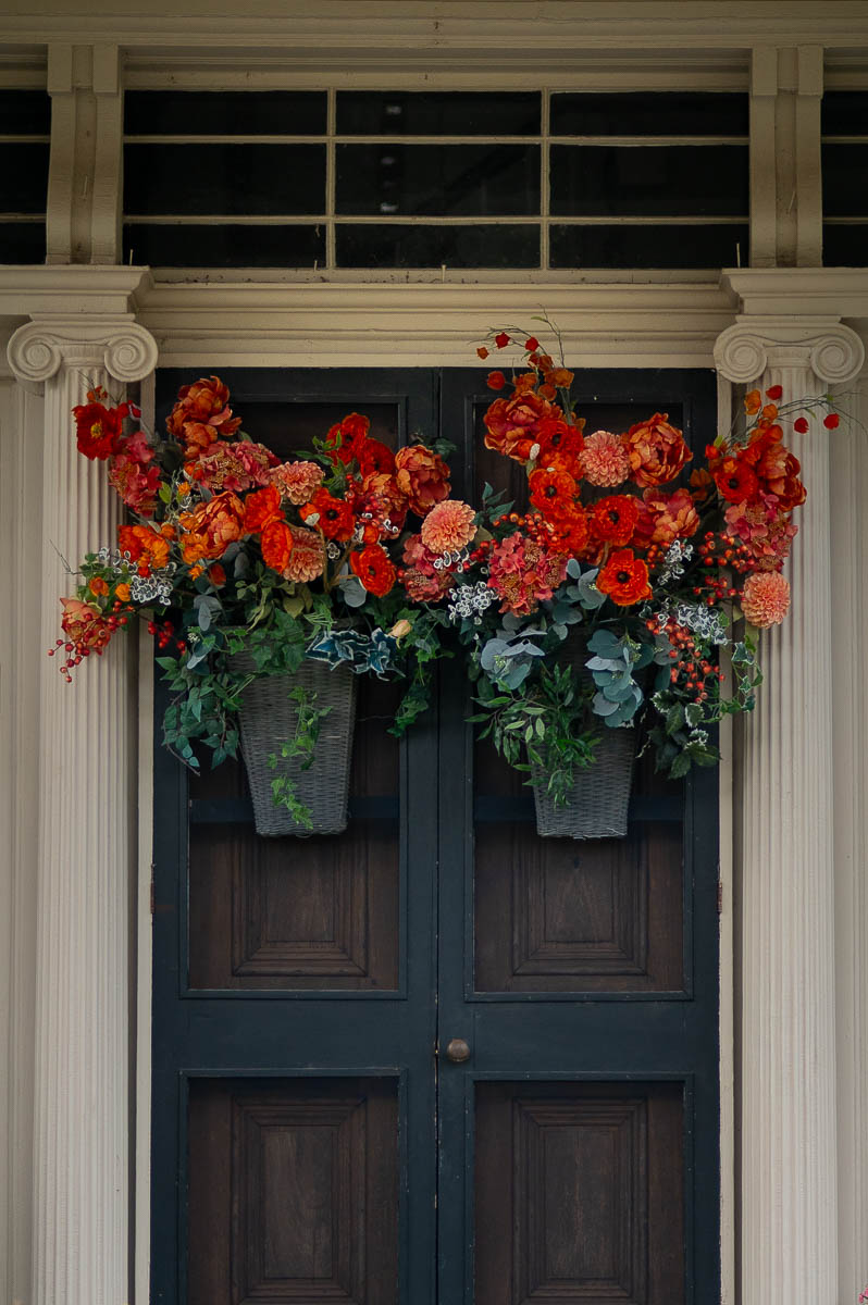 Instead. ofusing fall wreaths for front door, this fall front porch went about decorating their front door for autumn by hanging baskets filled with fall flowers on their door.