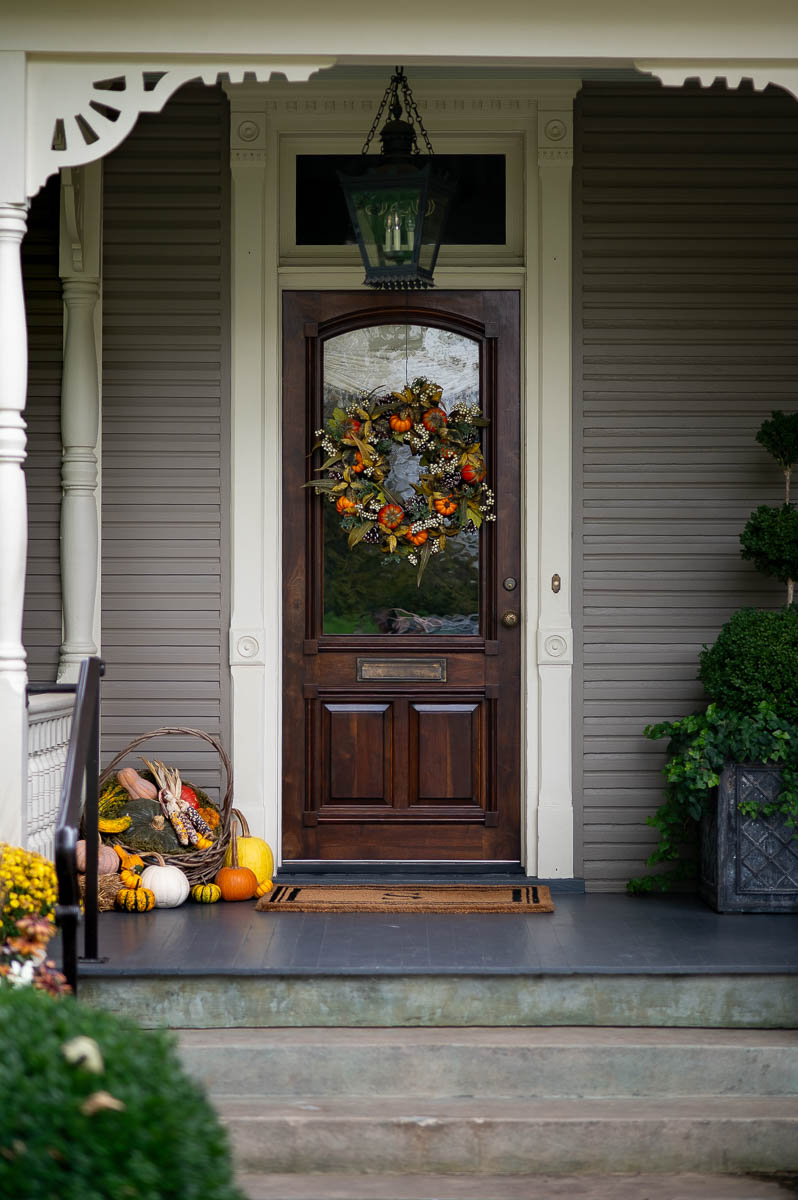 Elegant autumn front porch decor on a historic southern home featuring a fall wreath for front door and wooden basket filled with pumpkins, gourds, corn and squash.