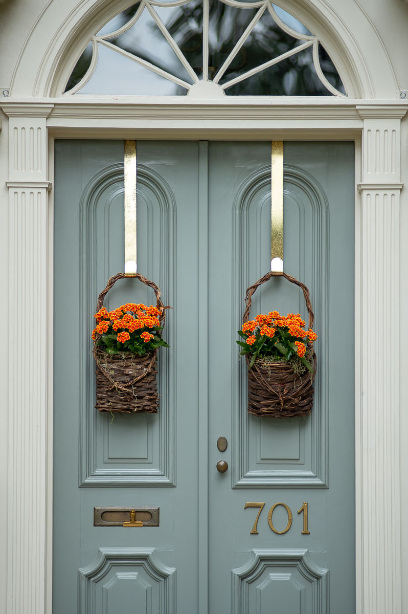 Historic southern fall front door decor with wicker hanging baskets filled with orange flowers hung from brass hooks in front of a blue door in Huntsville Alabama.