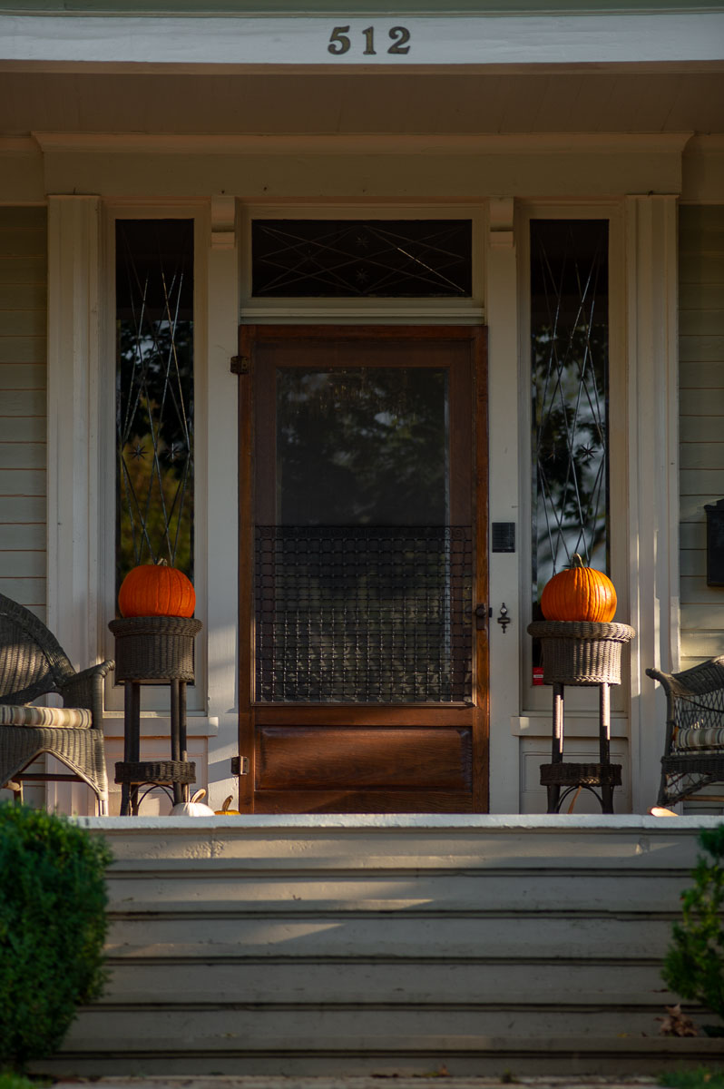 simple fall front porch idea with two pumpkins resting on wicker pedestals to either side of a front door on a historic home.