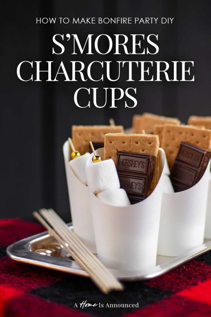 S'mores Charcuterie Cups filled with marshmallow skewers, chocolate bars and graham crackers sit on a silver tray on a red and black check blanket ready to serve at a backyard bonfire party.
