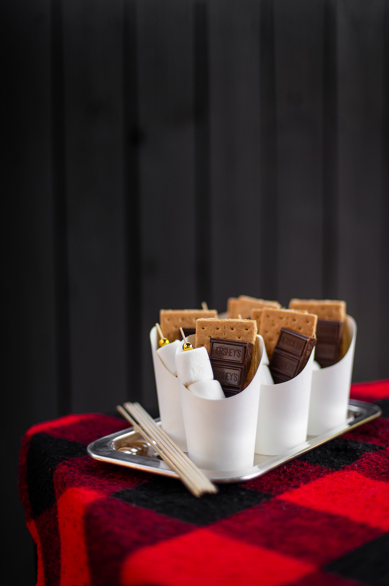 DIY S'mores Charcuterie Cups filled with marshmallow skewers, chocolate bars and graham crackers sit on a silver tray on a red and black flannel blanket ready to serve at a camping themed party