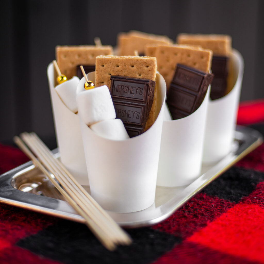 DIY S'more charcuterie cups filled with chocolate, marshmallow skewers and graham crackers on a silver try with bamboo roasting sticks for a bonfire party