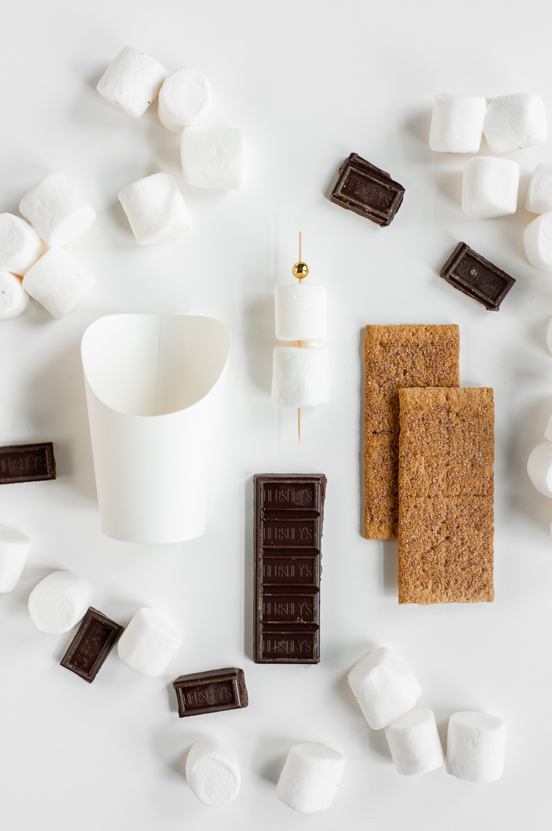 Ingredients arranged to assemble S’mores Charcuterie Cups with graham crackers, chocolate bars, and marshmallows on skewers.