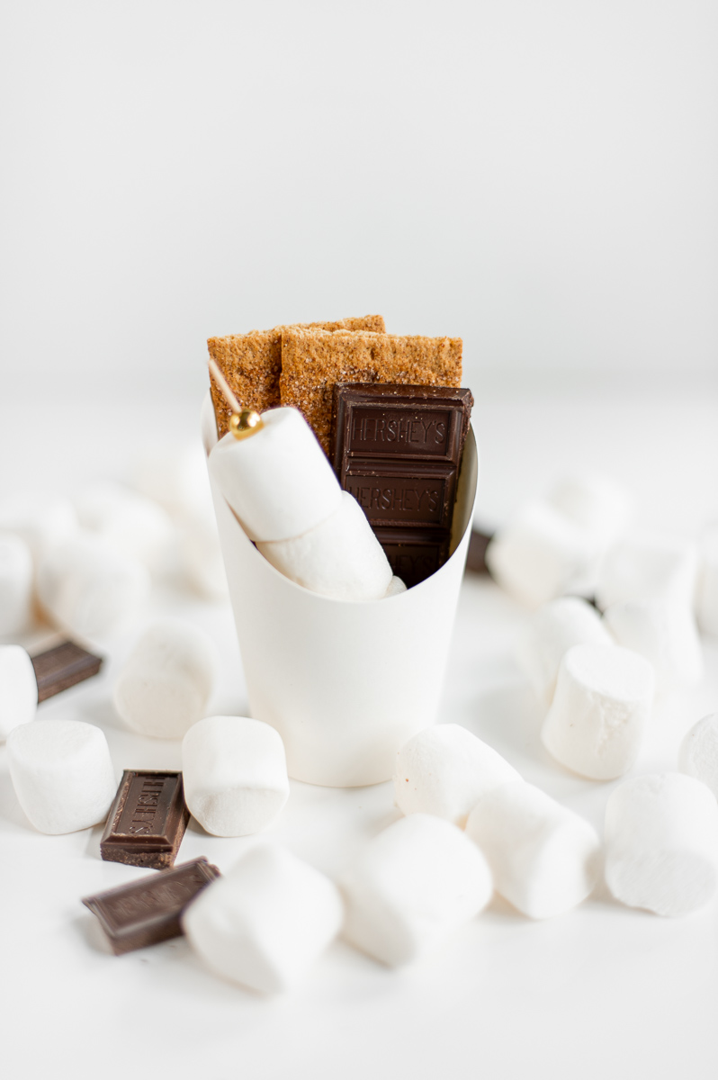 Assembled S’mores Charcuterie Cup on a white table with graham crackers, chocolate, and marshmallows on a skewer.