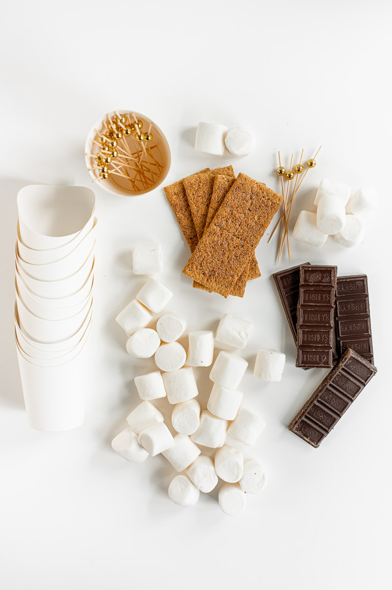 Overhead view of ingredients for S’mores Charcuterie Cups on a white table, including graham crackers, chocolate, and marshmallows.