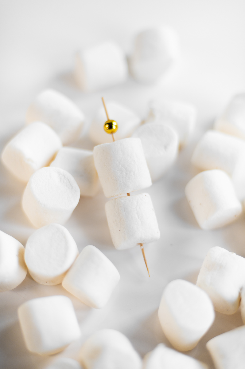 Two marshmallows on a skewer ready to place in a S’mores Charcuterie Cup with chocolate and graham crackers.