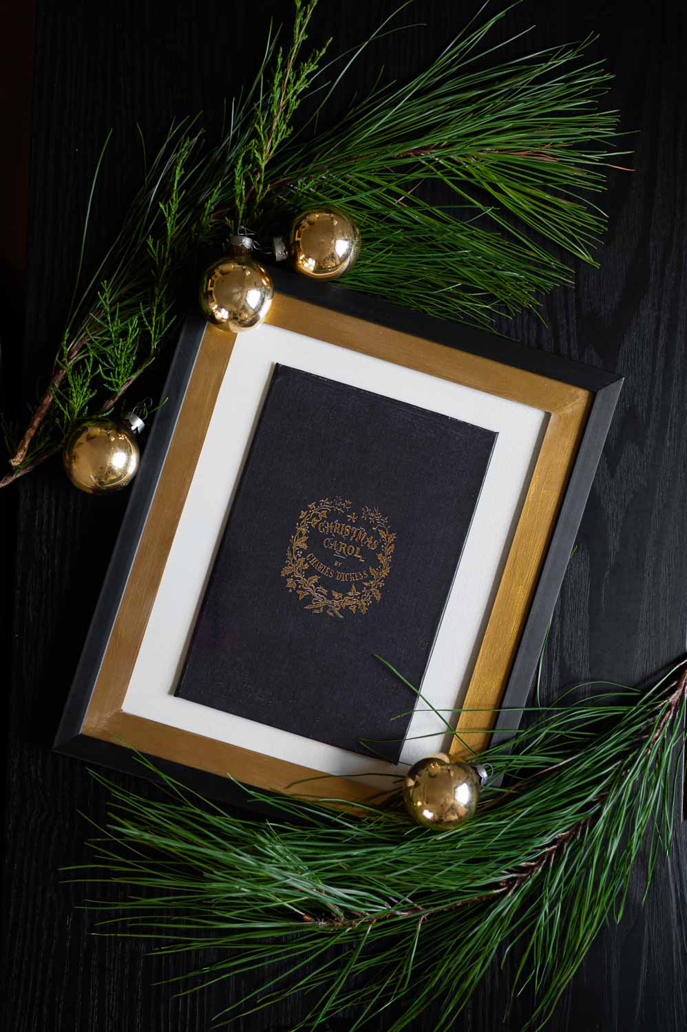a diy faux vintage book cover of a christmas carol is shown framed laying on a table surrounded by christmas garland and gold ornaments