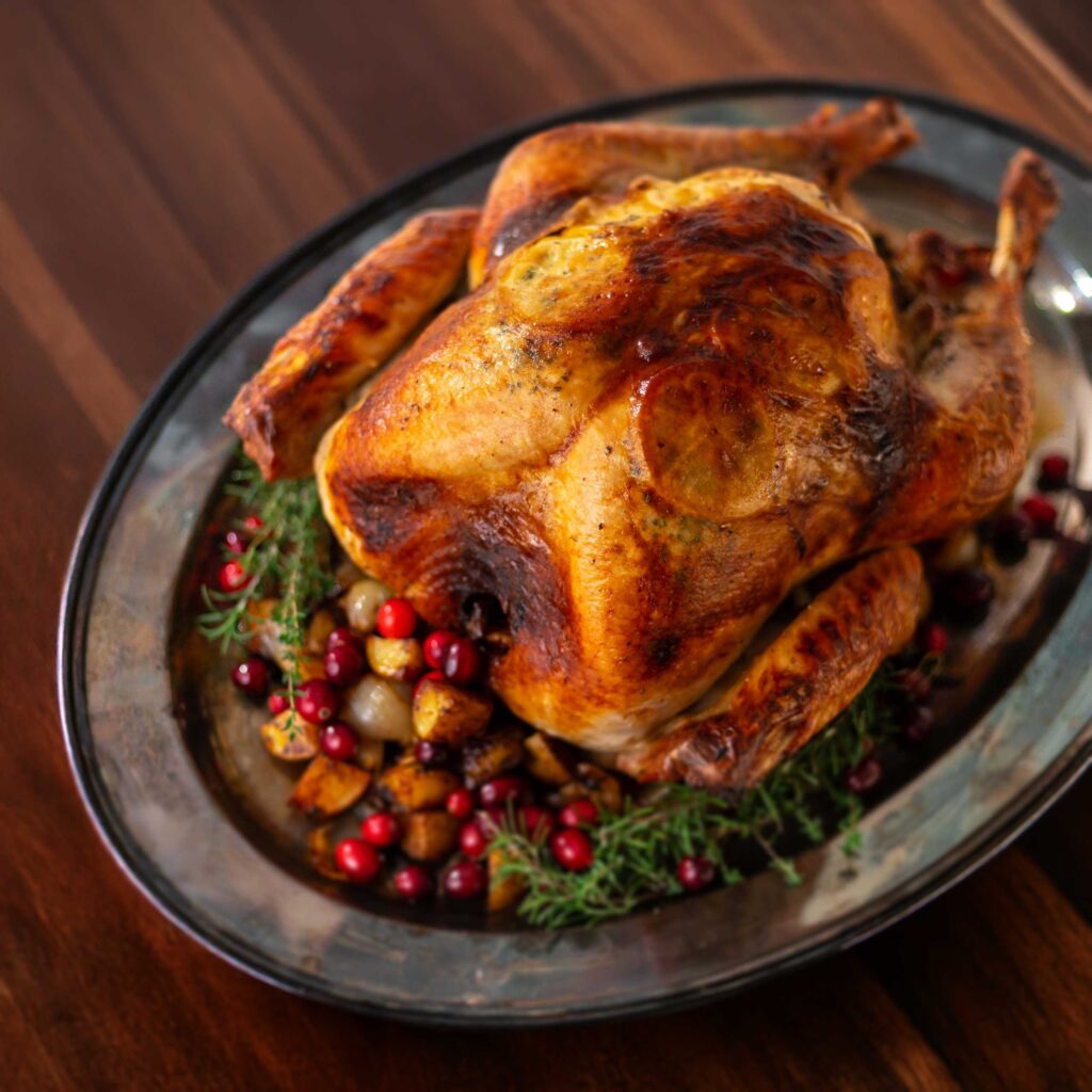 A classic thanksgiving dinner turkey rests on a silver tray surrounded by potatoes herbs and cranberries on a wood tabletop ready to be served.