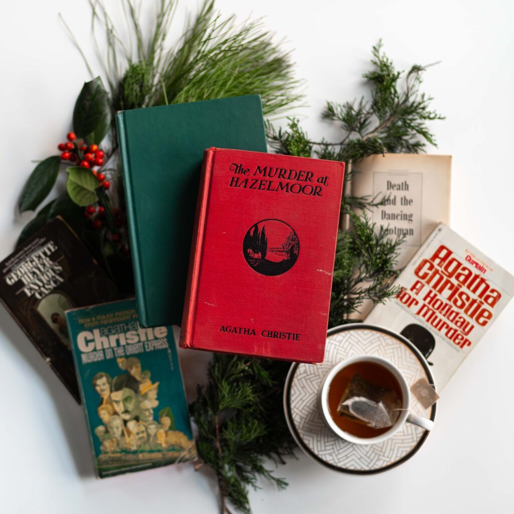 a collection of the best Christmas mystery books to read in winter written by golden age mystery writers laid out on a white table with evergreen branches next to a cup of tea