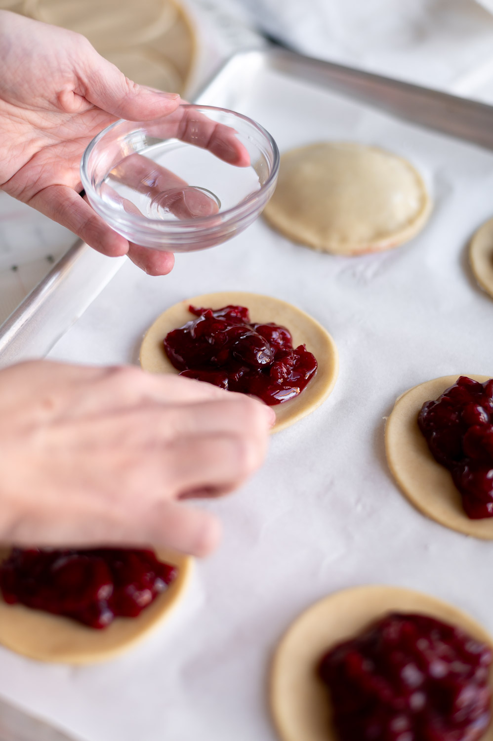 a woman uses her finger to wet the edges of pie dough before placing the lid on cherry hand pies to help seal the edges closed