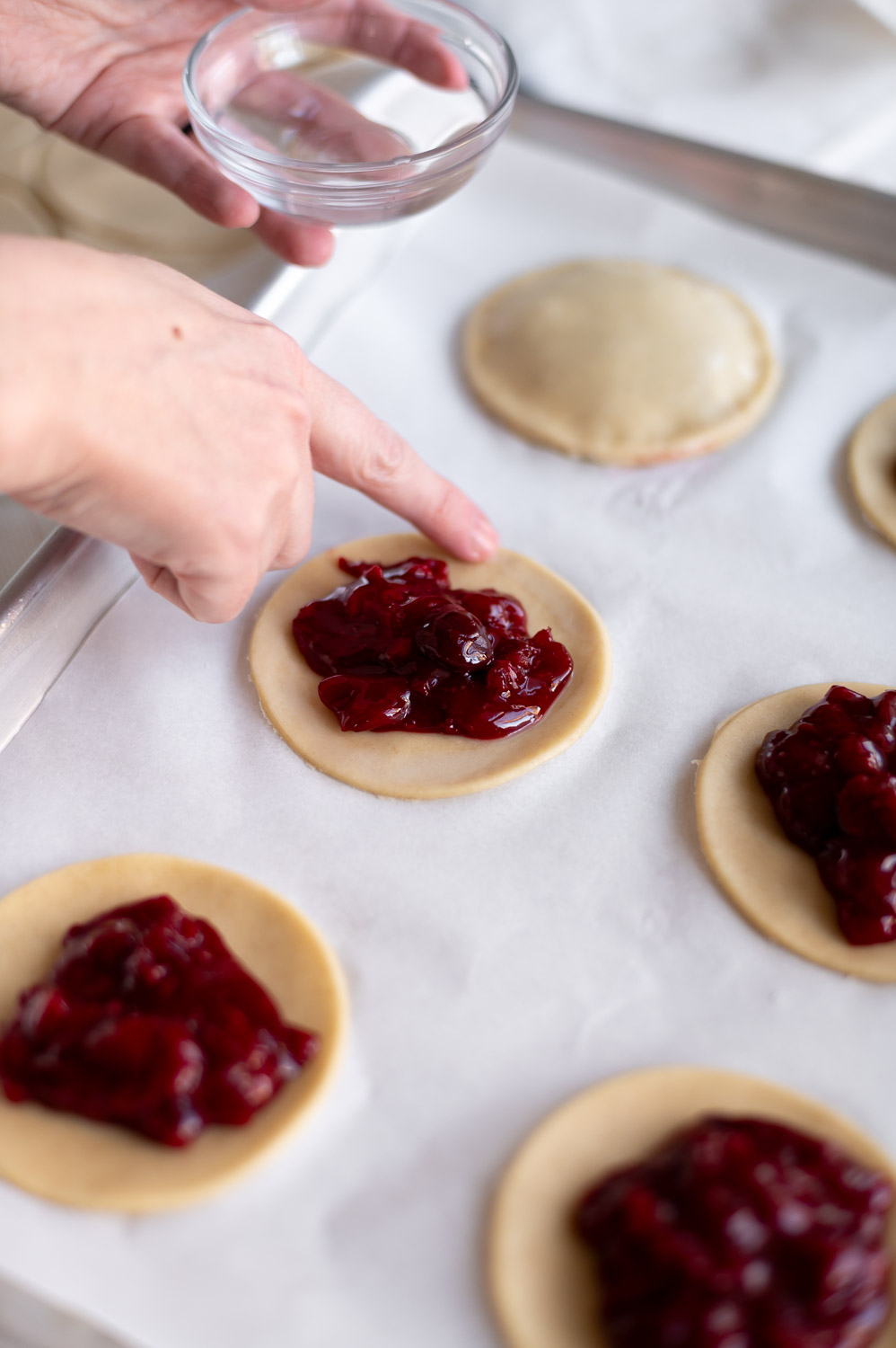 a woman uses her finger to wet the edges of pie dough before placing the lid on cherry hand pies to help seal the edges closed