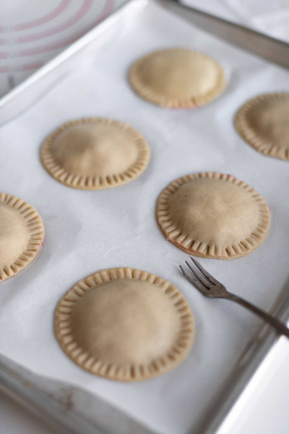 a tray of cherry hand pies rests on a table with their edges crimped closed by a fork ready to bake