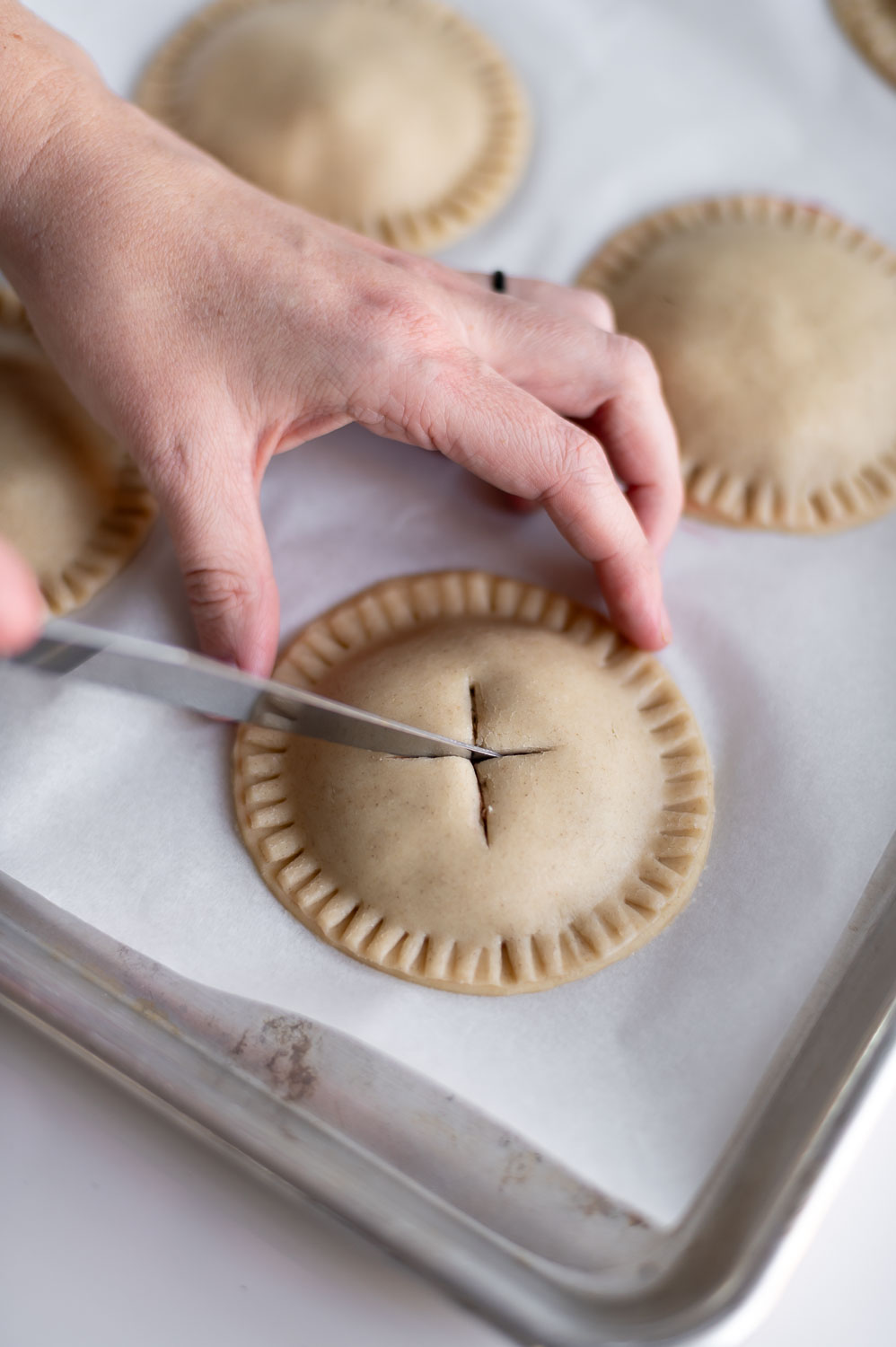 a woman gently cuts criss crossed vent holes in the same of an X on top of a handheld cherry pie before baking
