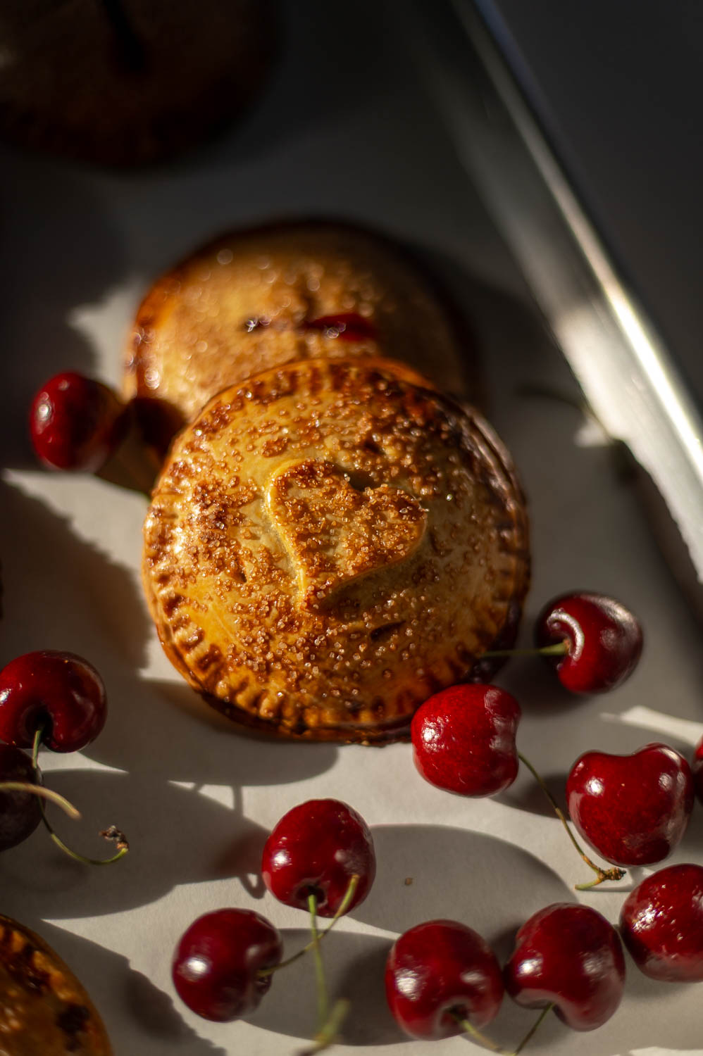 Valentine's day cherry hand pies fresh out of the oven rest on a baking sheet next to fresh cherries
