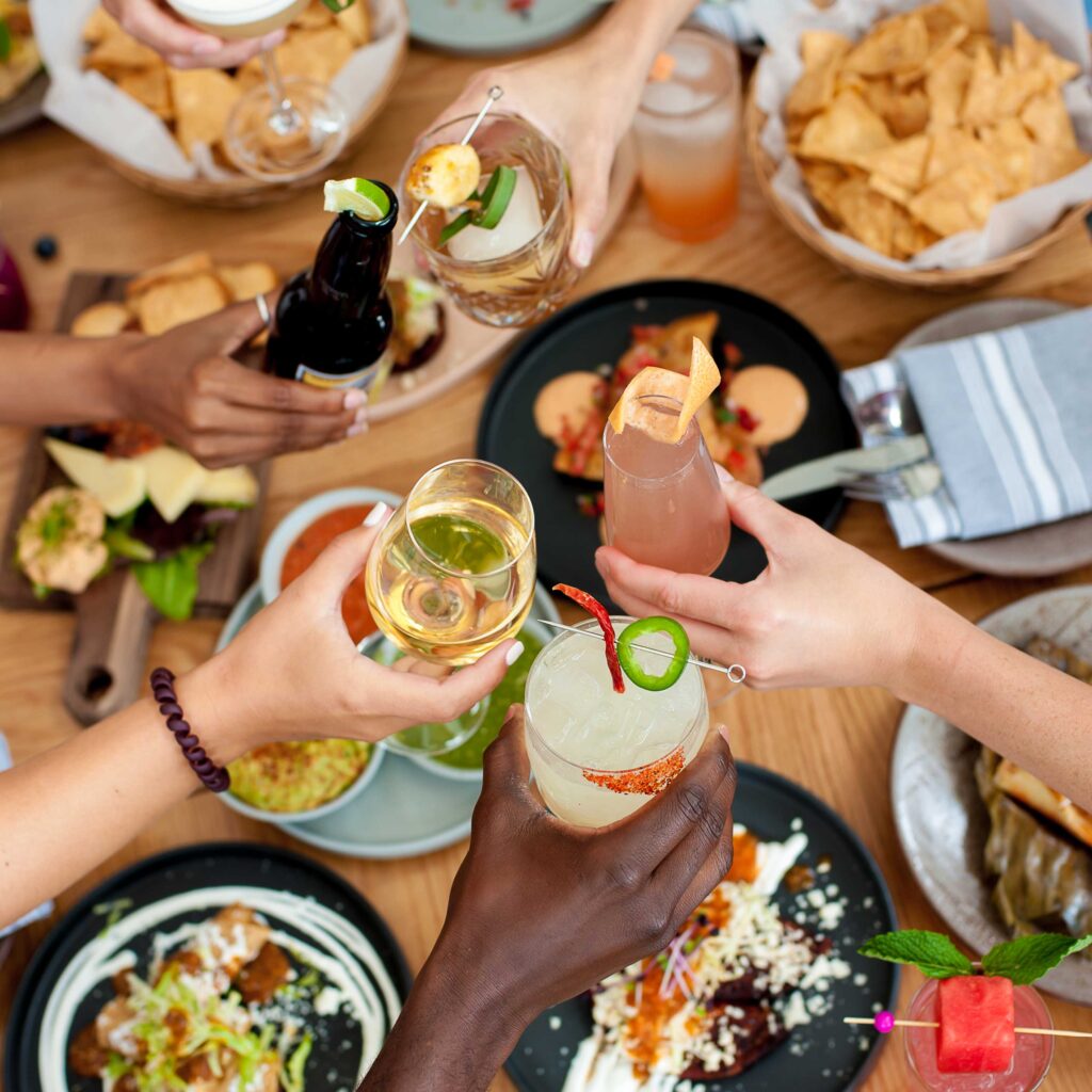 a group of people sitting around a table at a monthly supper club meeting for friends clicking glasses of various drinks together.
