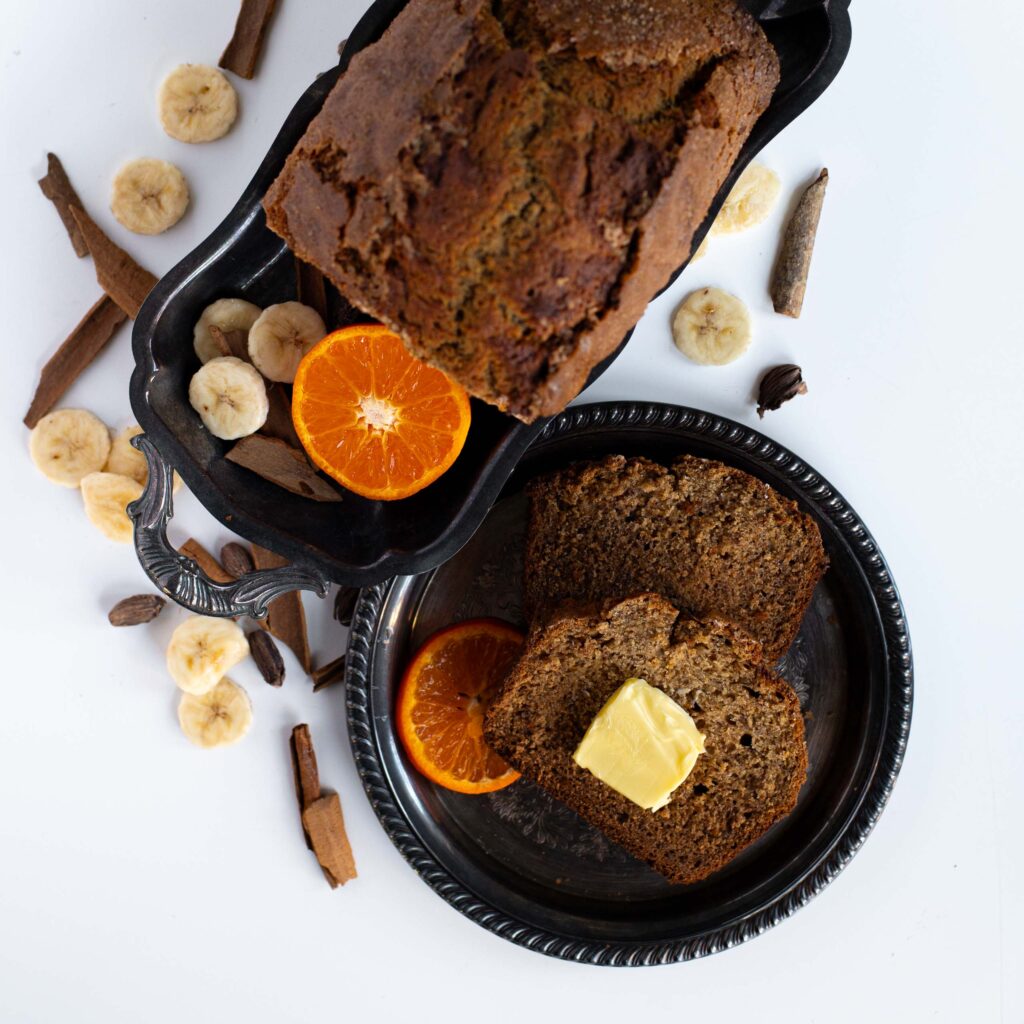 A loaf of orange cardamom brown butter banana bread rests on a silver tray next to two slices of banana bread displayed on a silver plate next to it topped with a pat of butter. Cardamom pods, slices of banana, clementines oranges and cinnamon sticks are scattered around the tabletop.