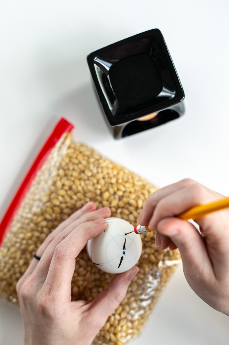 a woman makes a make with hot wax on an easter egg using the pin-drag drop pull wax resist method for decorating pysanky style Easter eggs without a kistka
