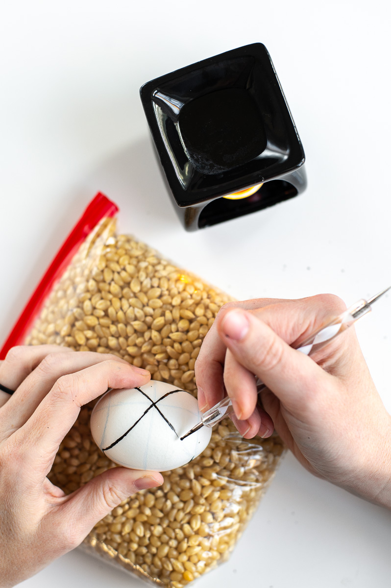 a woman draws on a easter egg with hot wax using an embossing stylus tool in order to create pysanky inspired easter egg designs without a kistka.
