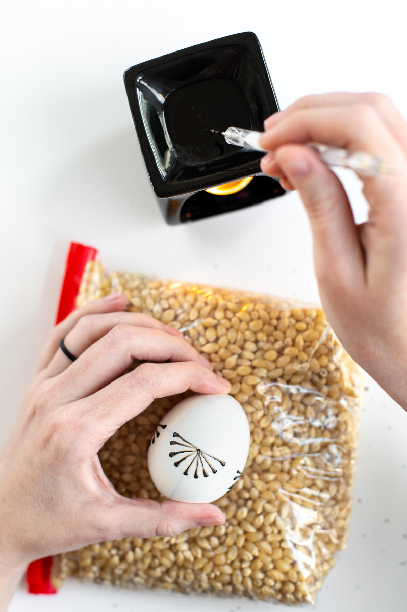 a woman dips a pin head in hot wax prior to using it to decorate an easter egg with crayon wax