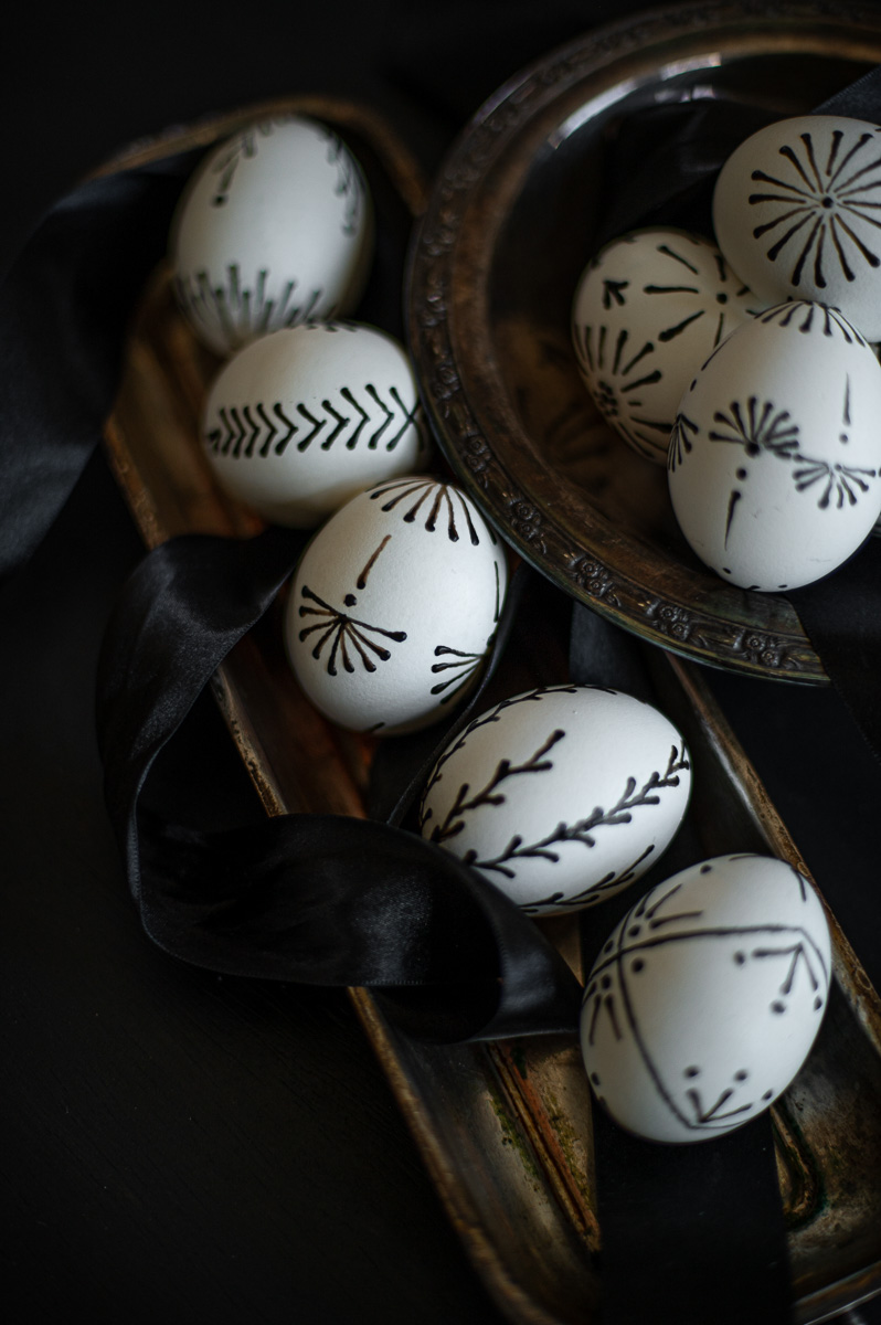 elegant black and white easter eggs with designs created out of wax rest in silver trays next to black satin ribbon.