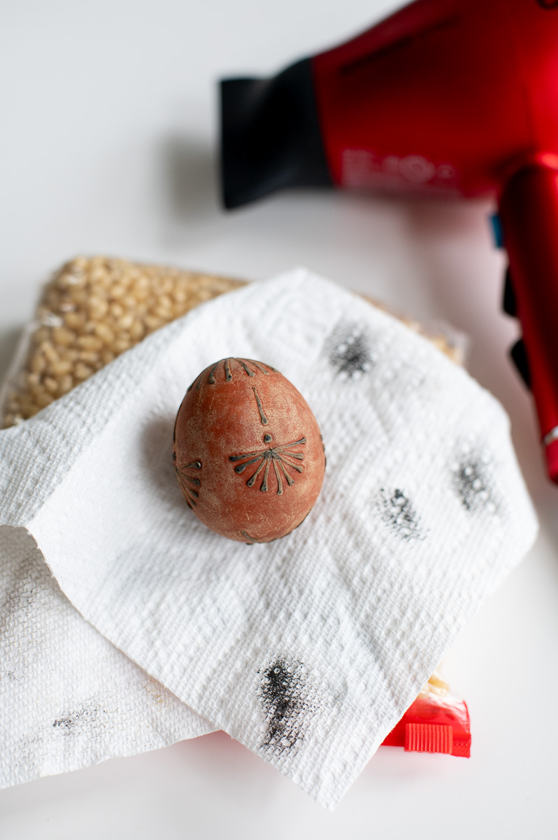 a woman removes wax of an easter egg decorated with crayon wax and dyed with onion skins using hot air from a blow dryer.