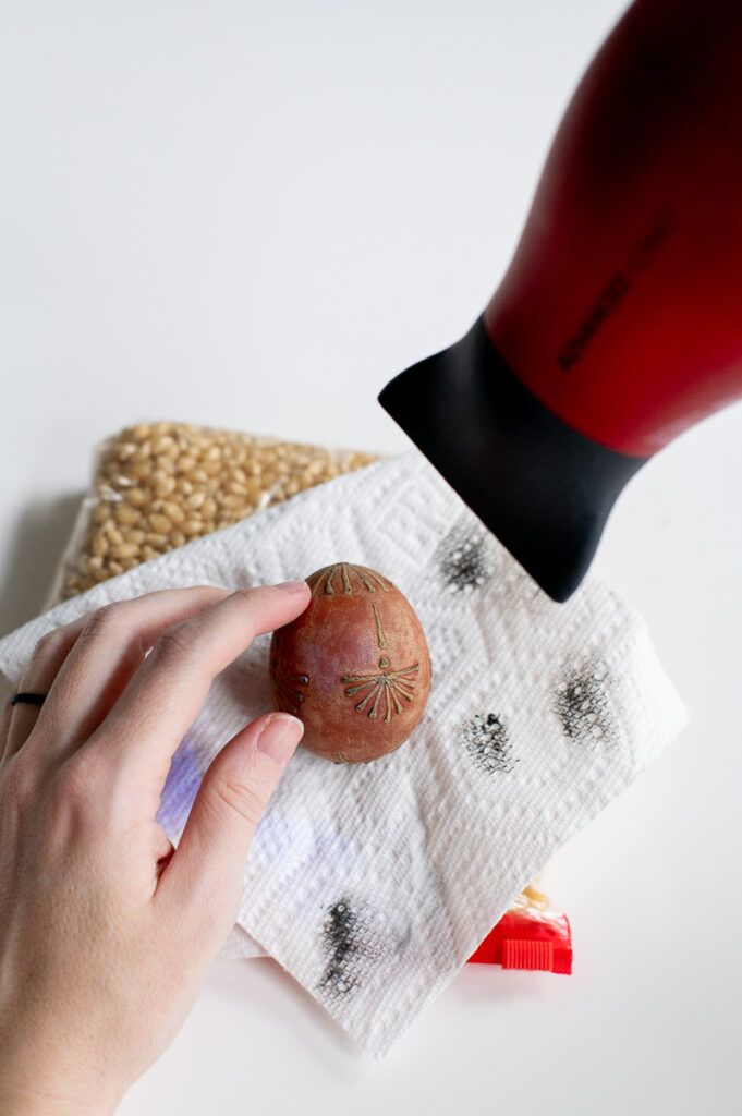 a woman remove wax of an easter egg decorated with crayon wax and dyed with onion skins using hot air from a blow dryer.