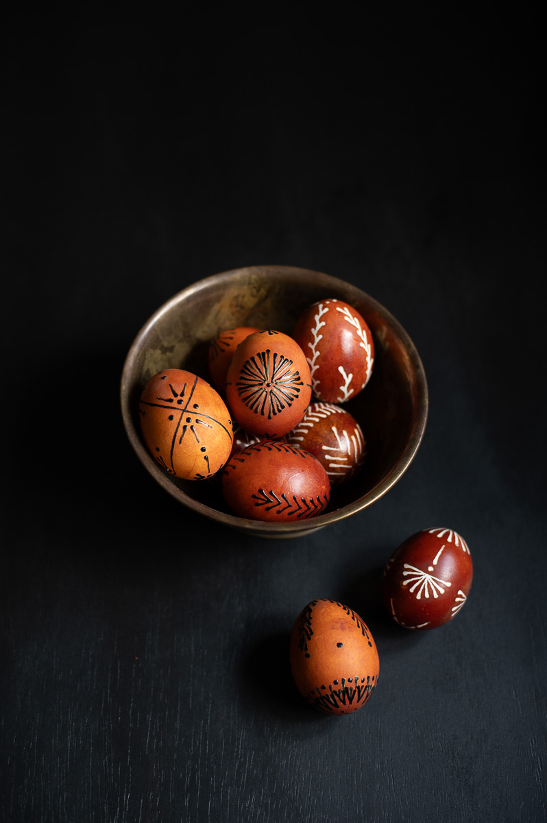 a brass bowl sits on a black table filled with wax resist easter eggs and pin-drag drop pull wax design eggs that have been dyed with onion skins.