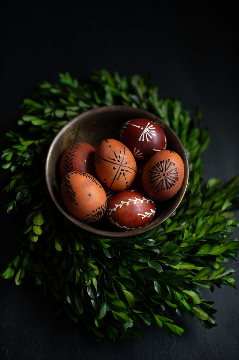 a bowl filled with traditional Pysanky easter eggs sits on a black table surrounded by boxwood branches as elegant Easter egg decor.